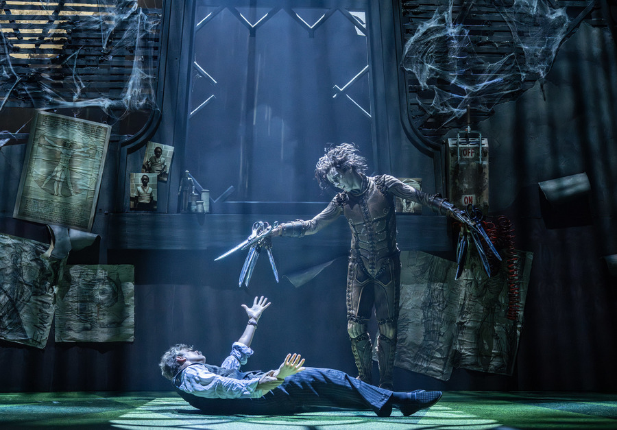 Photo from stage production of Edward Scissorhands. A man lies on the floor while Edward Scissorhands looms over him, waving his scissor hands.