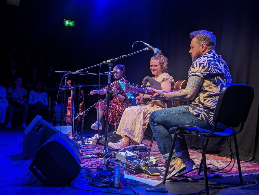 Photograph of Malin Lewis and band on stage at Tradfest. They sit on chairs holding their instruments.