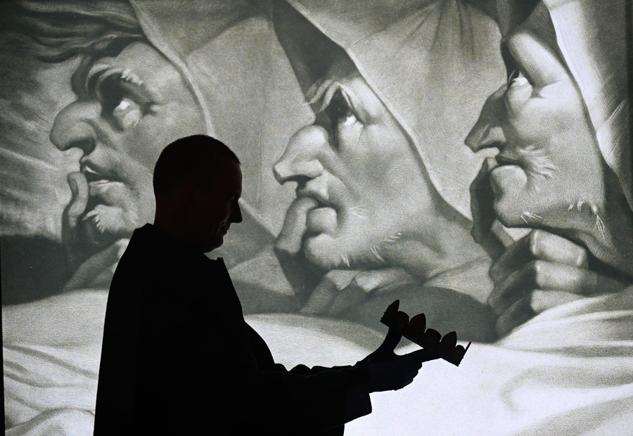 Black and white photo of a man holding a crown in front of a painting of three men in hoods.