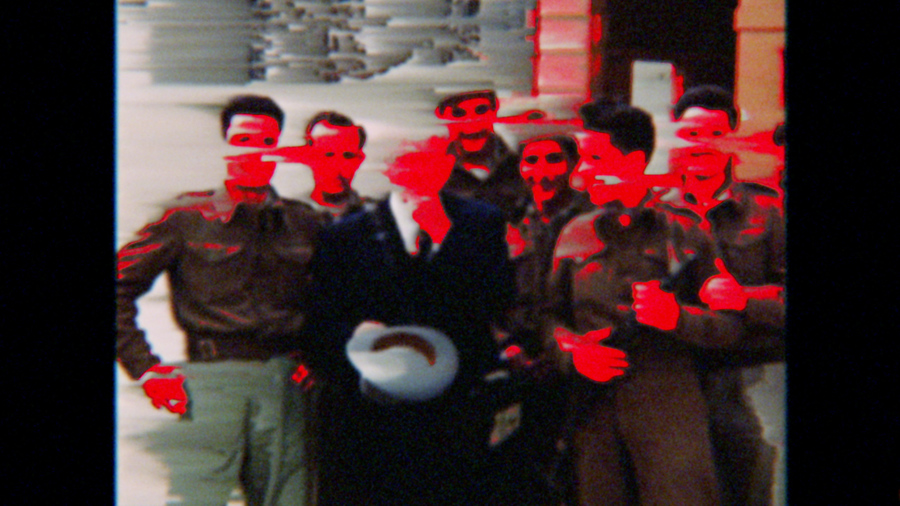 Film still depicting a group of men posing for a photo, with many of their faces daubed in bright red.