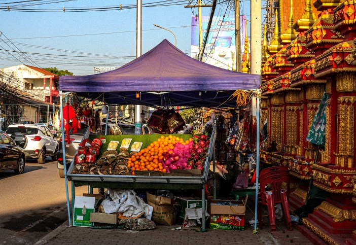 A photo of a fruit stall on the side of a road. The ornate walls of a temple are visible to the right, with cars passing on the left.