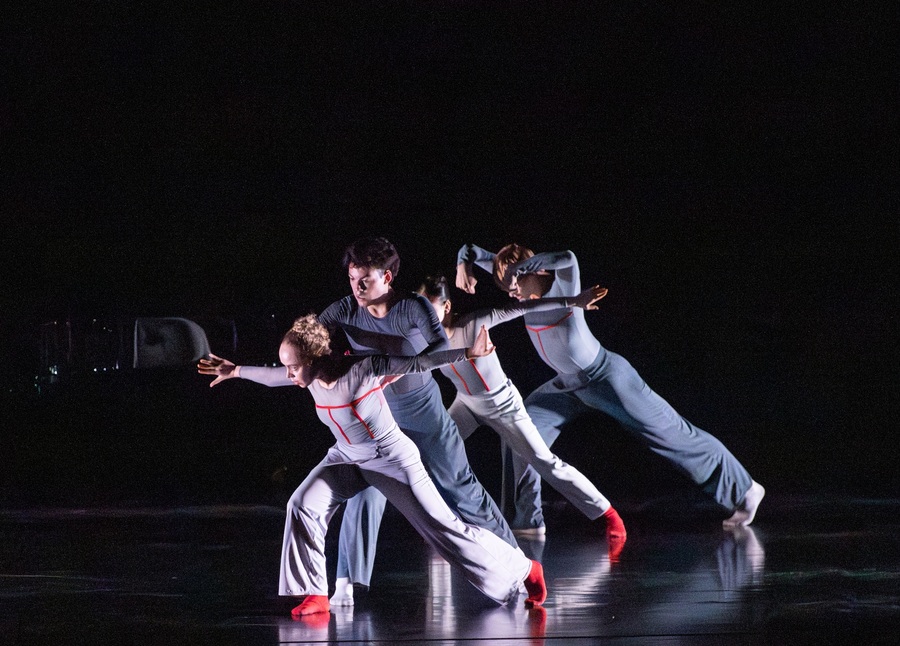 Four performers dance on stage in grey and red outfits.