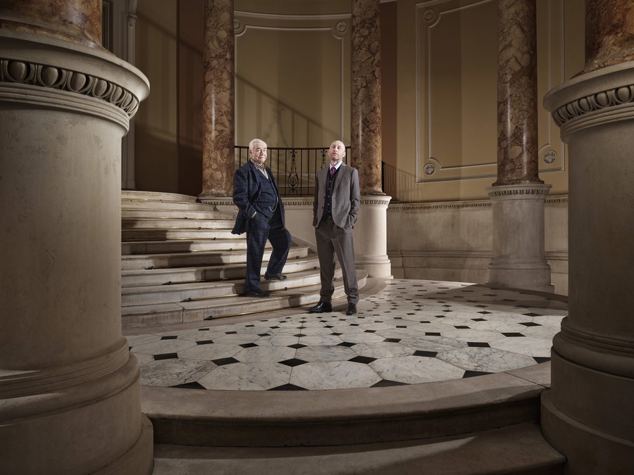 Brian Cox and Sandy Grierson stand at the bottom of a marble staircase.