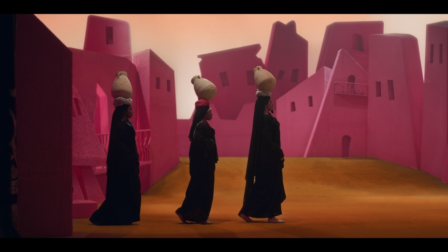 Film still showing three women in black robes, balancing water pots on their heads, walking past a row of bright pink buildings.