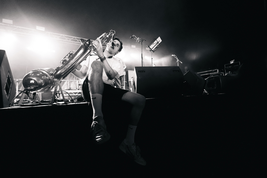 Black and white photo of a member of the band Black Foxxes sat on the edge of a stage playing the saxophone.
