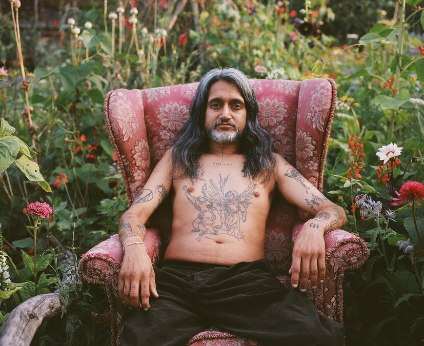 Photo of a long-haired male figure, sat in a red armchair among tall flowers and plants.