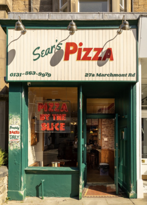 Photo of the exterior of Sears Pizza – a tenement shopfront painted in green, with a hand-painted sign above the doorway and a Pizza By The Slice neon sign in the window.