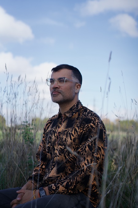 Photo of Brian D'Souza, wearing a brown patterned shirt, kneeling in a field of tall grass.
