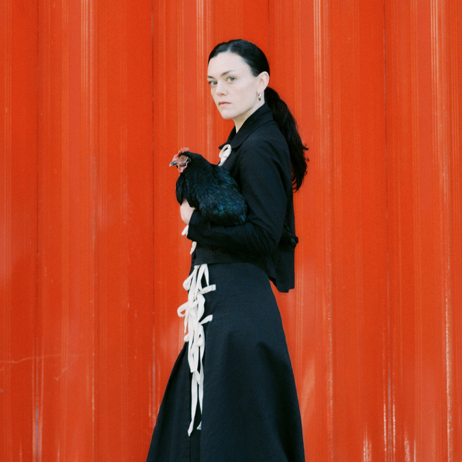 Album artwork for Iona Zajac's album Bang. Iona stands in front of a red wall, holding a chicken.