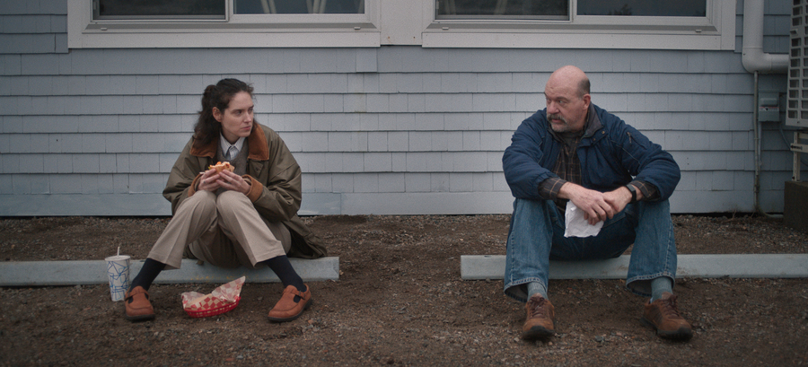 Still of Eva Victor and John Carroll Lynch in SORRY BABY; their characters sit side-by-side on a kerb, eating sandwiches.