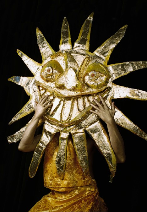Photo of a figure holding a large golden sun mask in front of their face.