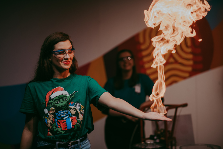 Photo of a woman in a green t-shirt conducting a science experiment in which she appears to be holding fire in her hand.