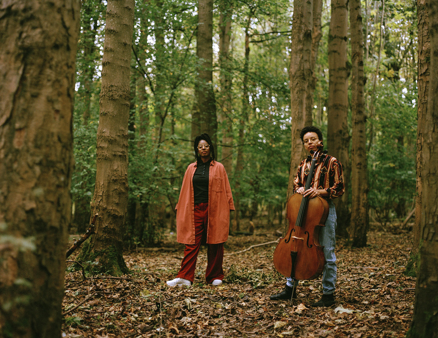 A photo of Mele Broomes and Simone Seales in a forest clearing, surrounded by trees. Simone holds their cello.