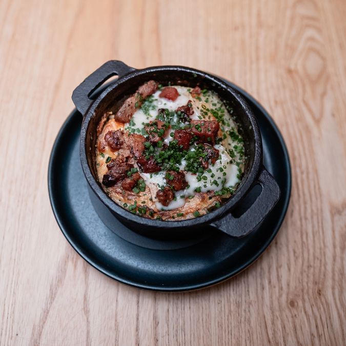 Photo of a small cast iron dish of tartiflette topped with bacon and herbs.