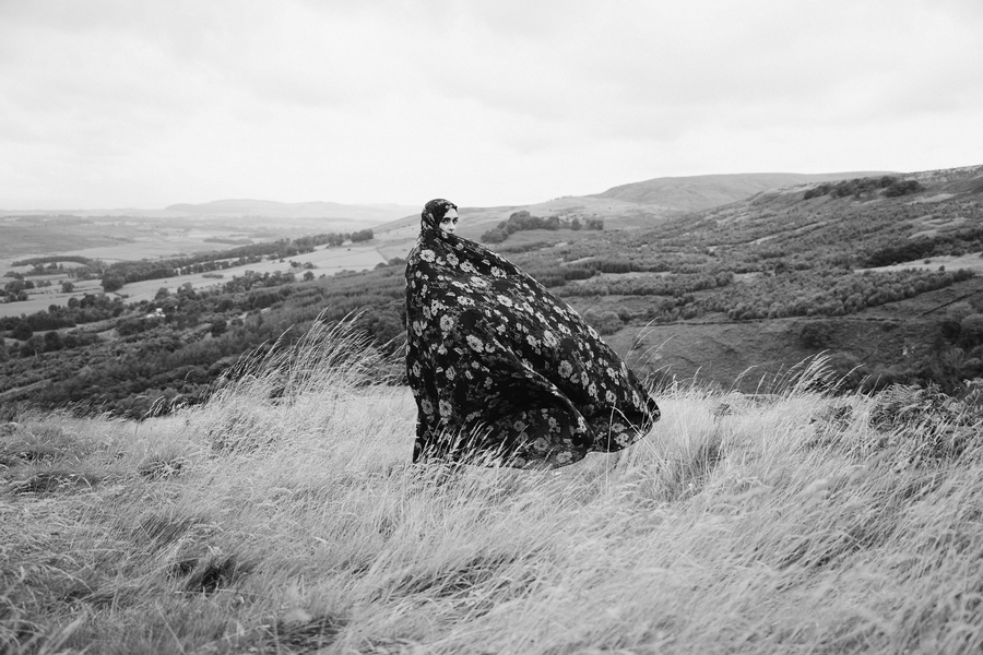 A black and white photograph of a woman in a long patterned robe and headscarf. The woman stands on a hill among long grass, with trees and fields visible in the background.