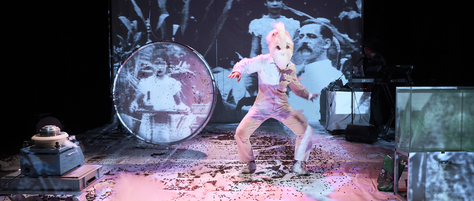 Photo of a figure in an all-white costume on stage, surrounded by objects and projected images.