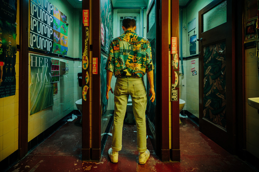 A still from Sleazy Tiger; a figure in a heavily patterned shirt stands in front of an open toilet cubicle