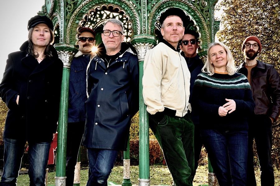 A photo of Belle and Sebastian, standing in a line by a Victorian park bandstand.