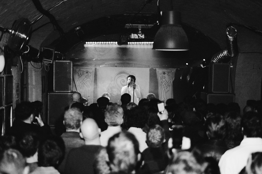 Black and white photo of a live performance by Madra Salach. Photo taken from the back of the crowd, with the backs of heads visible in front of the stage.