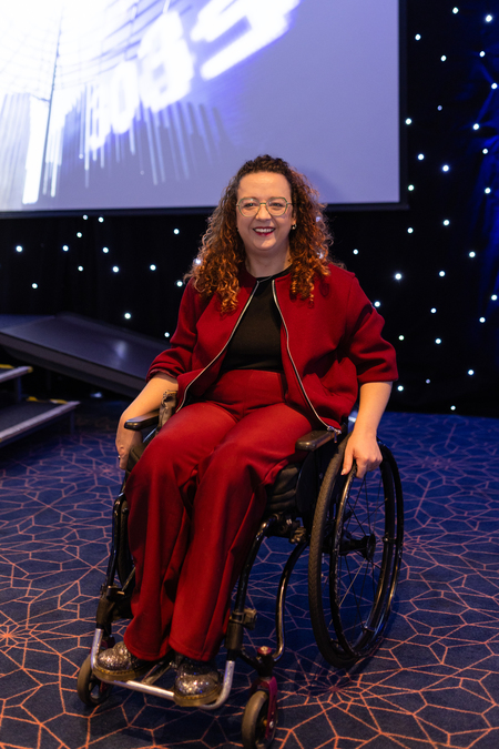 A photograph of Carrie-Ann Lightley. She wears a red zipped jacket and trousers, and sits in her wheelchair.