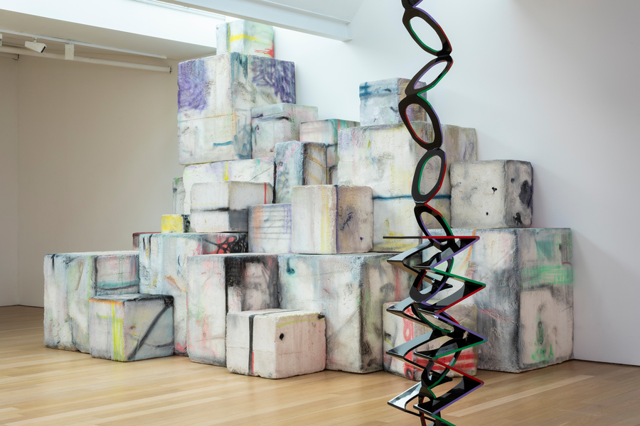 Photograph of two artworks in a gallery. At the back of the fram, a large structure made up of white and off-white cubes; in front, a thin geometric sculpture.